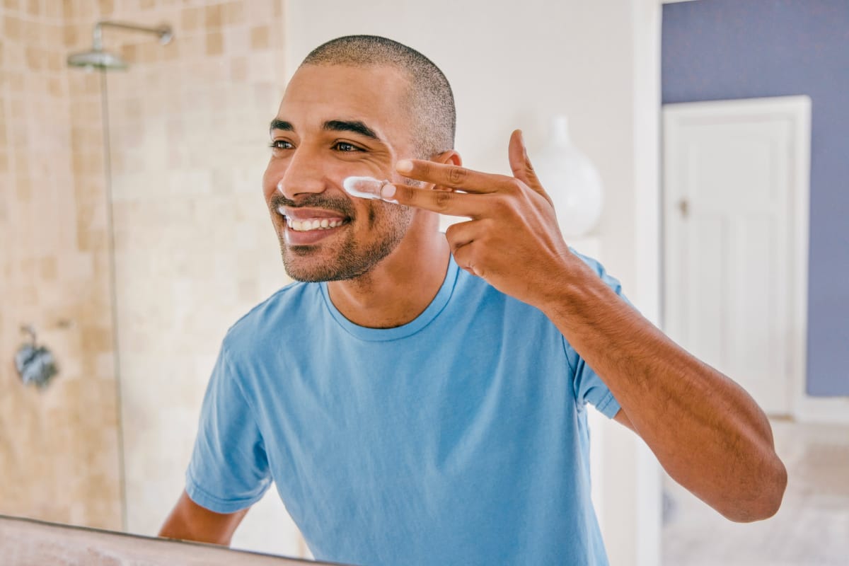 Millenial man smiling and applying morning skincare routine in bathroom mirror